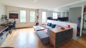 A spacious living room with a sofa, a coffee table, a desk with a computer, and a kitchen area visible through an open door.