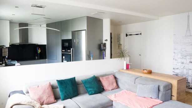 A modern living room with a grey sectional sofa, a wooden coffee table, and a large mirror reflecting the kitchen area.