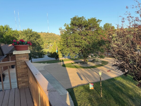 A residential area with a paved driveway, a brick wall, a wooden deck, and a garden with a potted plant and a flag. - Hub to Black Hills, 1 acre in city, 2 King beds, 3BR, 2 1/2 BA,dbl heated garage (Rapid City)