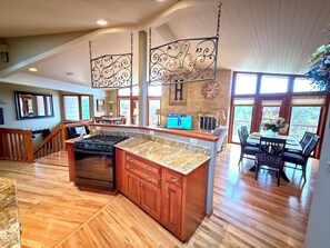 A spacious kitchen with a granite countertop, wooden cabinets, and a large island. There is a dining area with a table and chairs, and a view of the outdoors through large windows. - Hub to Black Hills, 1 acre in city, 2 King beds, 3BR, 2 1/2 BA,dbl heated garage (Rapid City)
