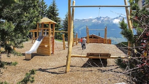 A playground with a slide and swings, surrounded by trees and a mountain view. - Lovely apartment for 4 guests with WIFI, pool and balcony (Les Collons)