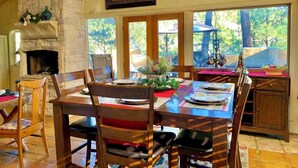 A dining area with a stone fireplace, wooden chairs, a table set for four, and a view of the outdoors through large windows.
