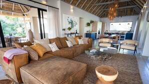 Ein geräumiger Wohnbereich mit einem großen braunen Sofa, einem Glastisch und einem gewebten Teppich. Auf dem Teppich steht eine Holzschale, und der Raum hat eine strohgedeckte Decke.
