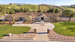 Une grande maison avec une allée incurvée, entourée d’une pelouse bien entretenue et d’un vignoble en arrière-plan.