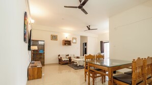 A dining area with a glass-top table and wooden chairs, a ceiling fan, and a wall-mounted TV.