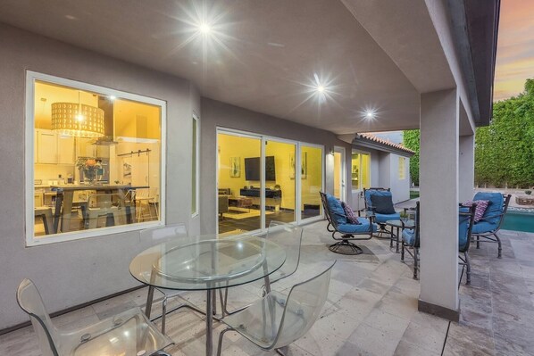 A modern patio with a glass table and chairs, a pool, and a well-lit interior with sliding glass doors. - Meadow Hills in North Scottsdale / Heated Pool (Scottsdale)