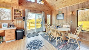 Interior de uma cabana de madeira com uma mesa de jantar, cadeiras, um ventilador de teto e grandes janelas que oferecem vista para o exterior.