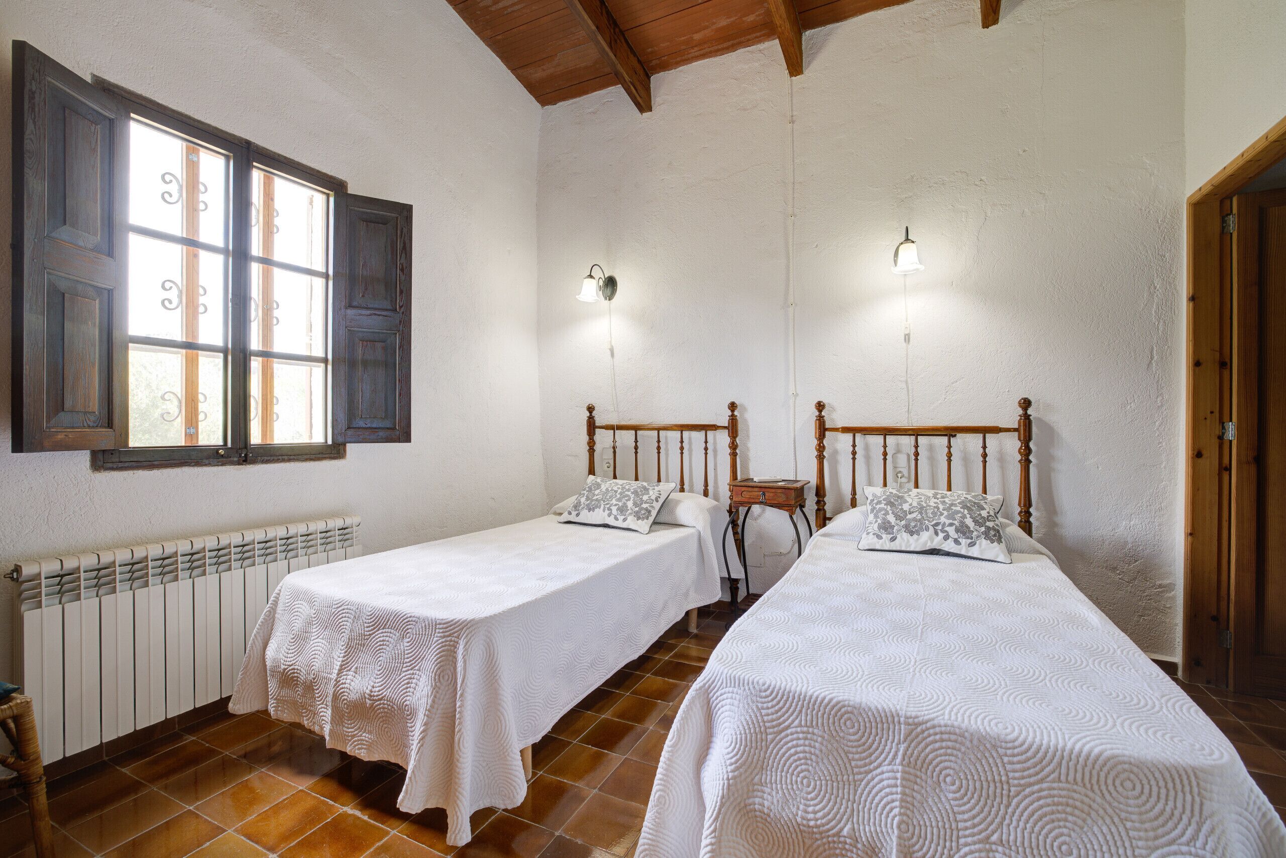 A room with two single beds, a radiator, a window with shutters, and a wooden ceiling.
