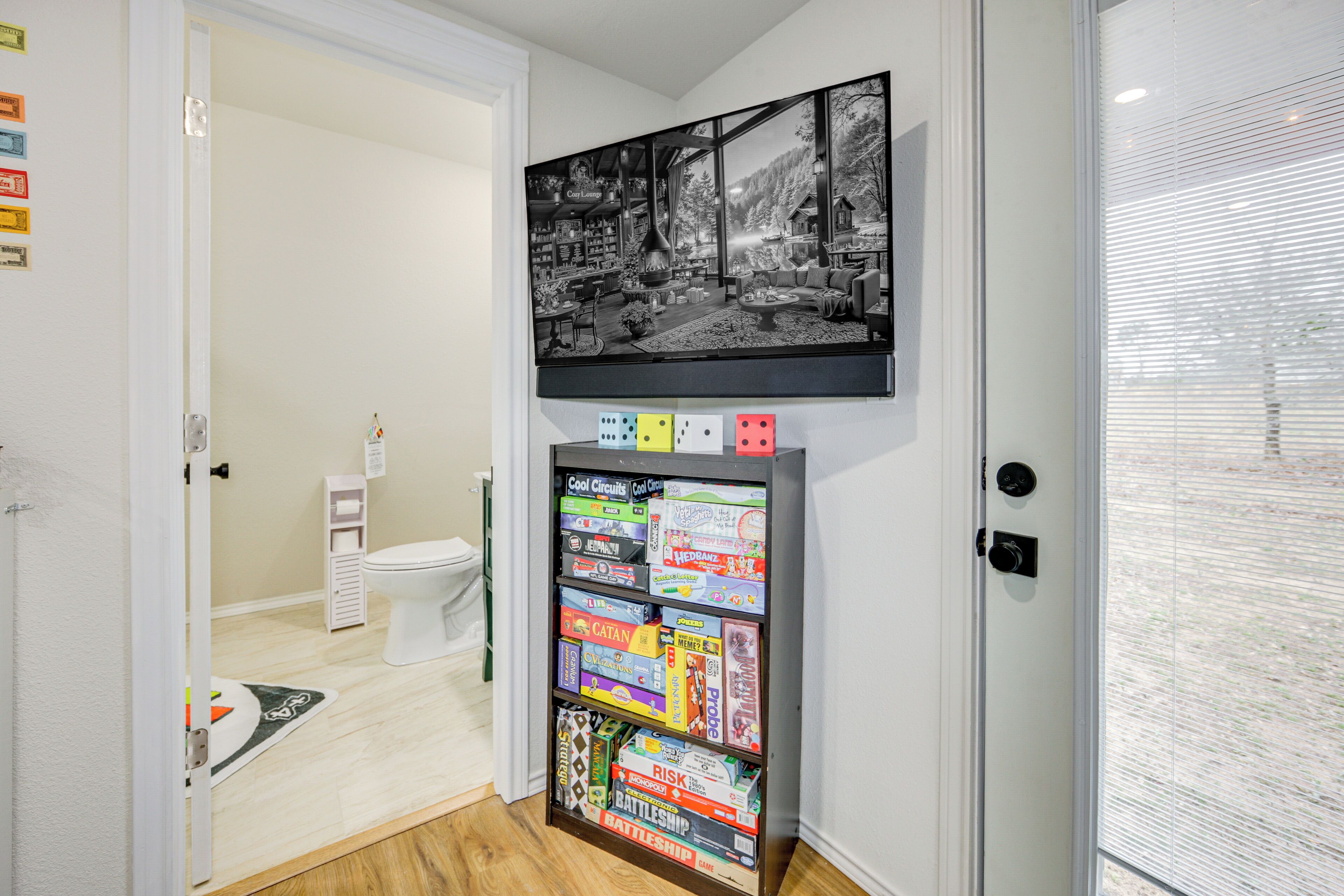 A bathroom with a toilet, a sink, and a mirror. There is a shelf with various board games and a framed picture above the shelf.