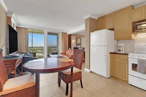 A modern hotel room with a balcony, a dining area with wicker furniture, a kitchenette with a refrigerator and stove, and a flat-screen TV.