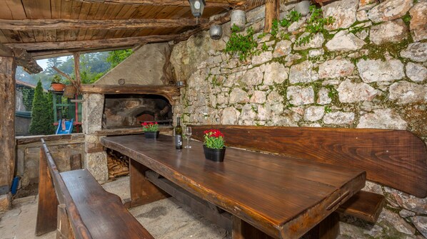 A rustic stone wall with a wooden table and benches, a stone oven, and a view of greenery outside.