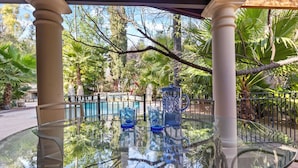 A glass table with chairs and a decorative pitcher, surrounded by a pool and palm trees.
