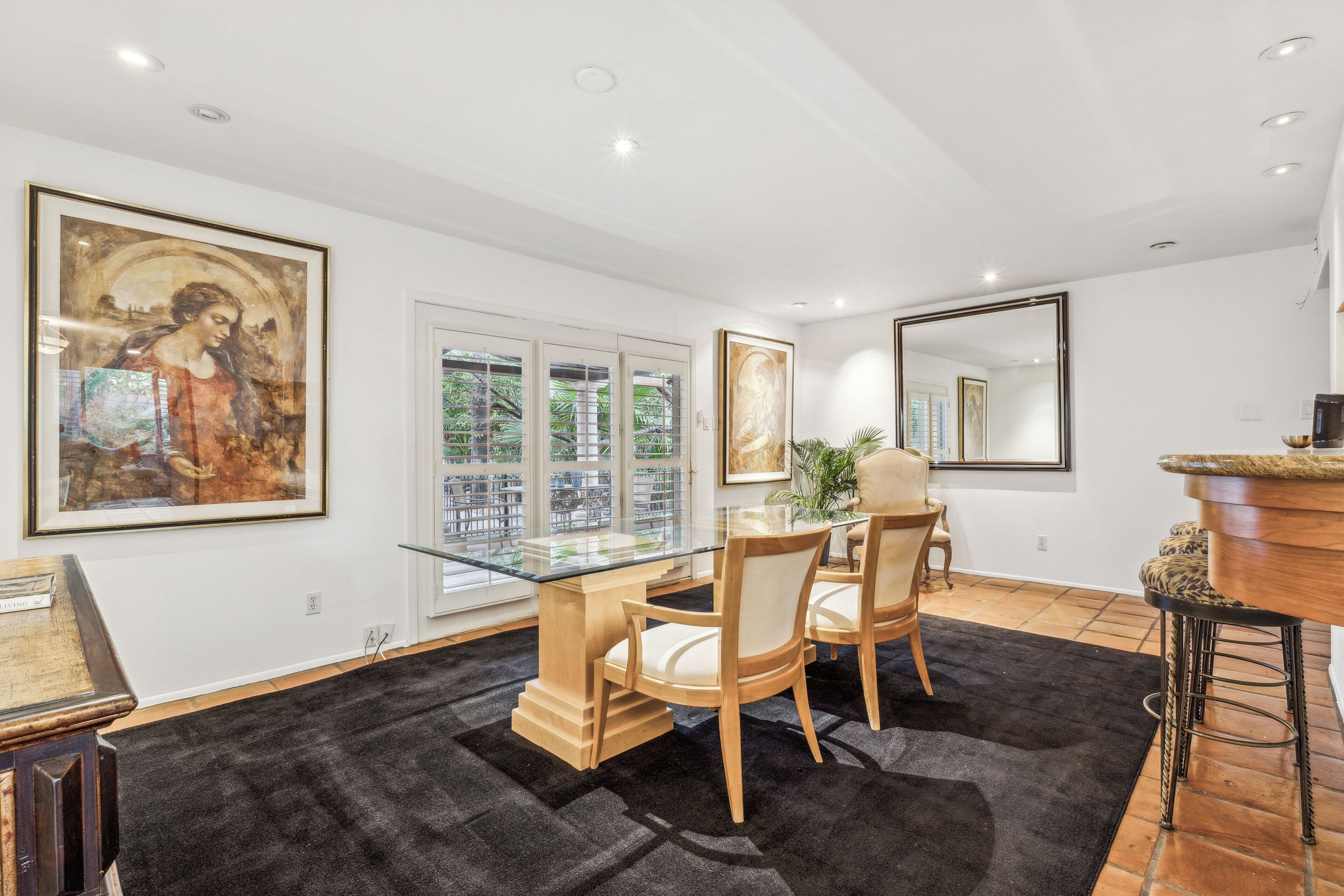 A dining area with a glass-top table, wooden chairs, and a black rug. There are framed artworks on the walls and a large mirror.