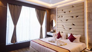 Quarto de hotel moderno com uma cama grande, piso de madeira, teto com iluminação embutida e uma janela com cortinas.