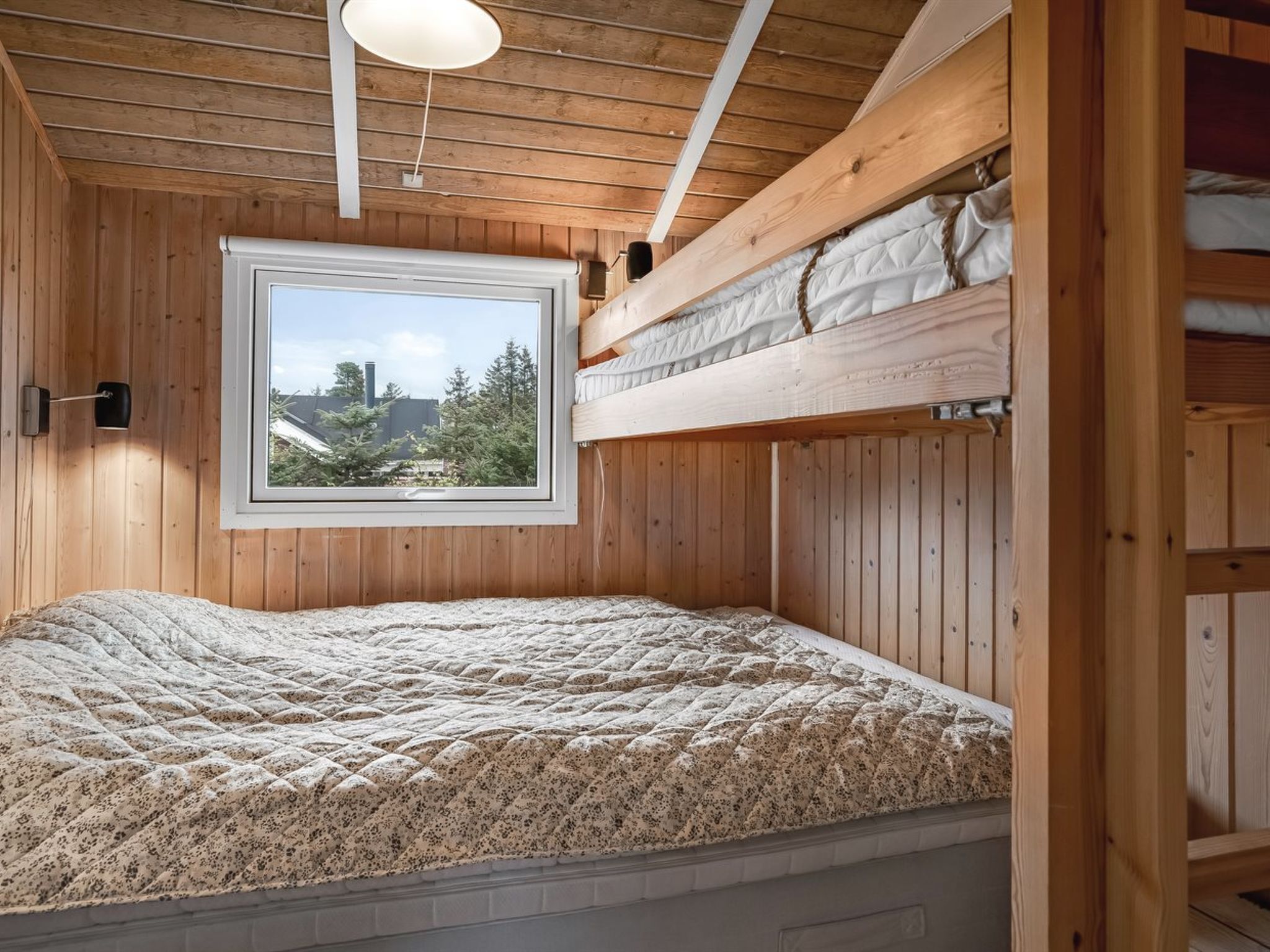 A wooden room with a bunk bed, a window with a view of trees and buildings, and a wall-mounted lamp.