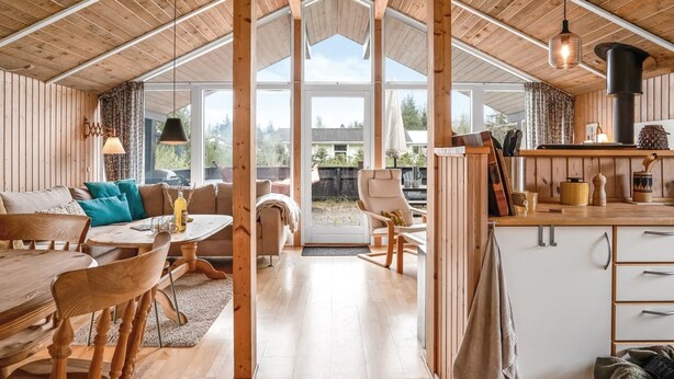 A spacious cabin interior with a high ceiling, wooden beams, a dining area, a kitchen, and large windows offering a view of the outdoors.