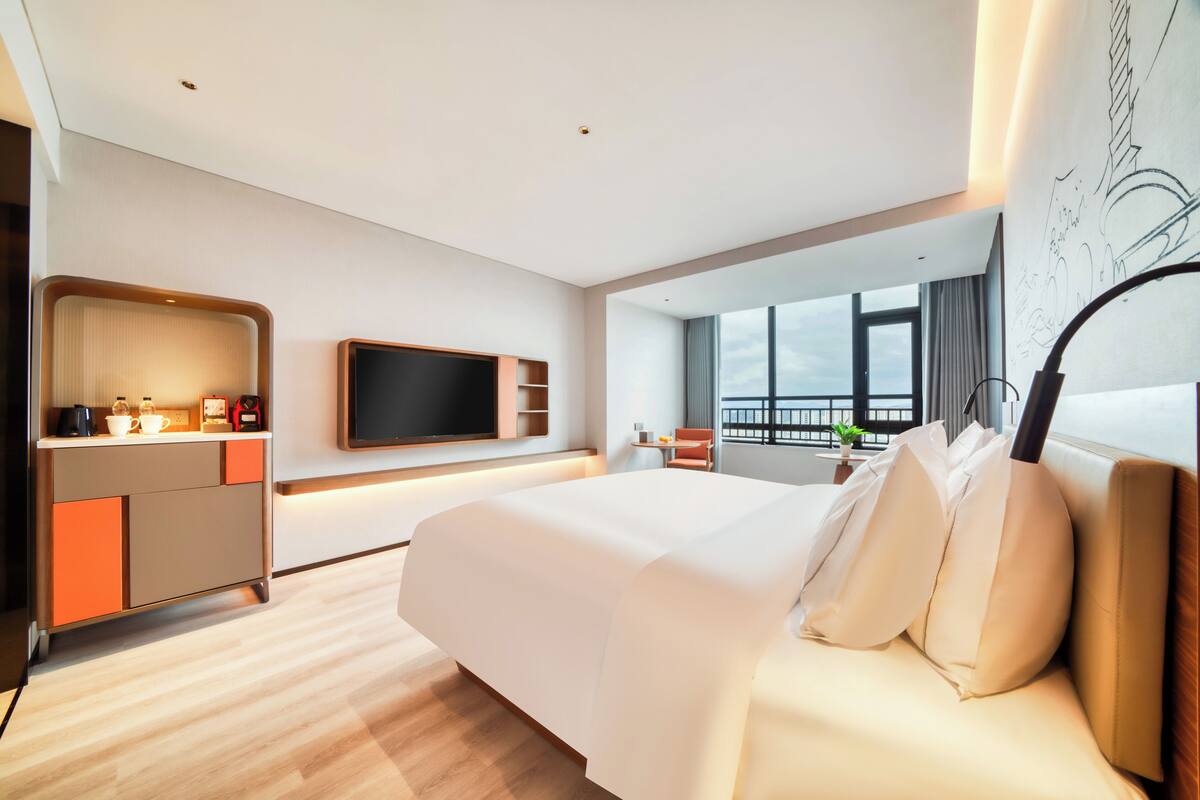 Ein modernes Hotelzimmer mit einem großen Bett, einem Flachbildfernseher, einem Schreibtisch mit Kaffeemaschine und Blick auf die Stadt durch große Fenster.