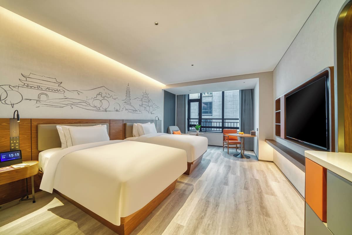 Ein Hotelzimmer mit zwei Betten, einem Schreibtisch mit einer Uhr, einem Fernseher und einem großen Fenster mit Blick auf die Stadt.