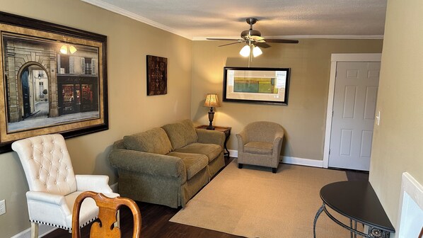 A living room with a sofa, armchair, and a small table with a lamp. There is a ceiling fan, a framed picture on the wall, and a door in the background. - The Monroe: 2-bedroom townhome. Walk to LSU (Baton Rouge)