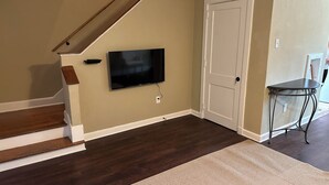 A staircase with dark wood steps and white trim, a flat-screen TV mounted on the wall, a white door, and a small table with a metal frame. - The Monroe: 2-bedroom townhome. Walk to LSU (Baton Rouge)
