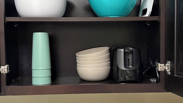 A kitchen with a dark wooden cabinet containing a white bowl, a teal colander, a stack of beige bowls, a teal container, and a black toaster. - The Monroe: 2-bedroom townhome. Walk to LSU (Baton Rouge)