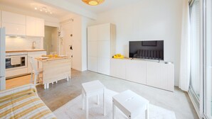 A modern, minimalist living room with a flat-screen TV, a dining area with a wooden table and chairs, and a kitchen with white cabinetry.