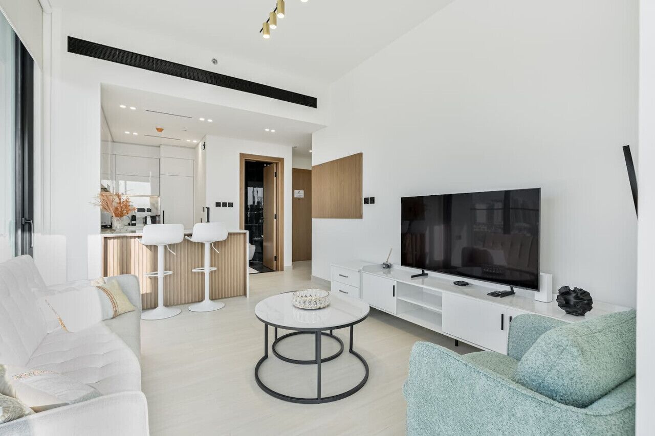 Una sala de estar moderna con un sofá, una mesa de centro y un televisor. Se observa una cocina al fondo.