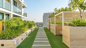 Un jardin sur le toit, avec un chemin central, entouré de jardinières remplies de verdure, et des bâtiments modernes en arrière-plan.