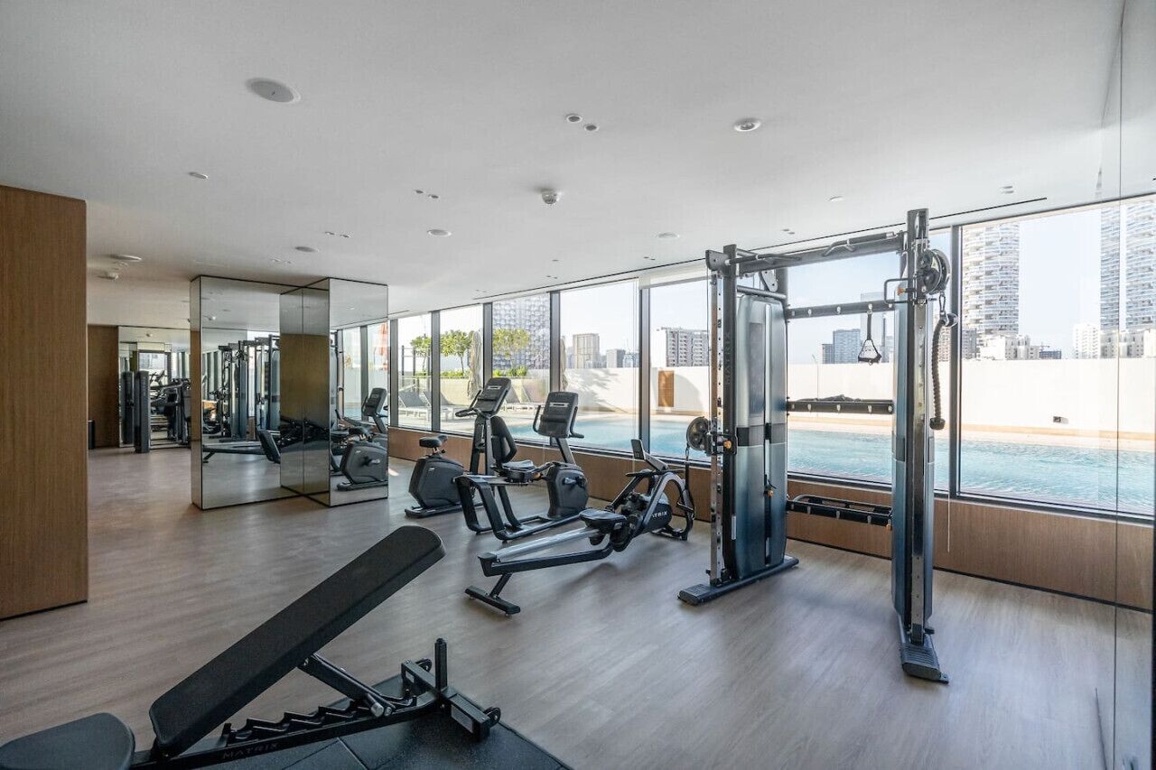 Un gimnasio moderno con máquinas de cardio, equipo de levantamiento de pesas y grandes ventanales que ofrecen vistas de la ciudad.