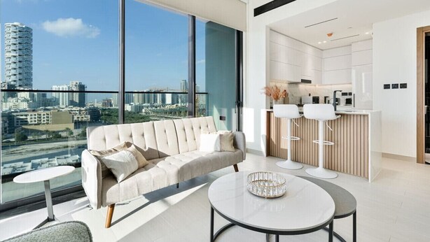 Un salon moderne doté d’une grande fenêtre, d’un canapé, d’une table basse et d’un espace cuisine visible à travers la vitre.