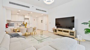A modern living room with a large flat-screen TV, a glass coffee table, and a sofa. There is a dining area with a table and chairs, and a kitchen in the background.