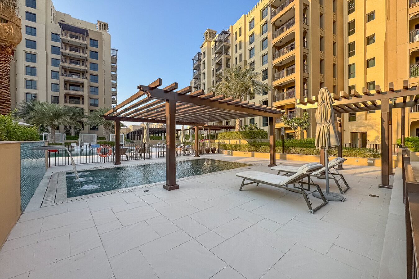 Uma área de piscina no terraço com espreguiçadeiras e uma pérgula, cercada por edifícios altos.
