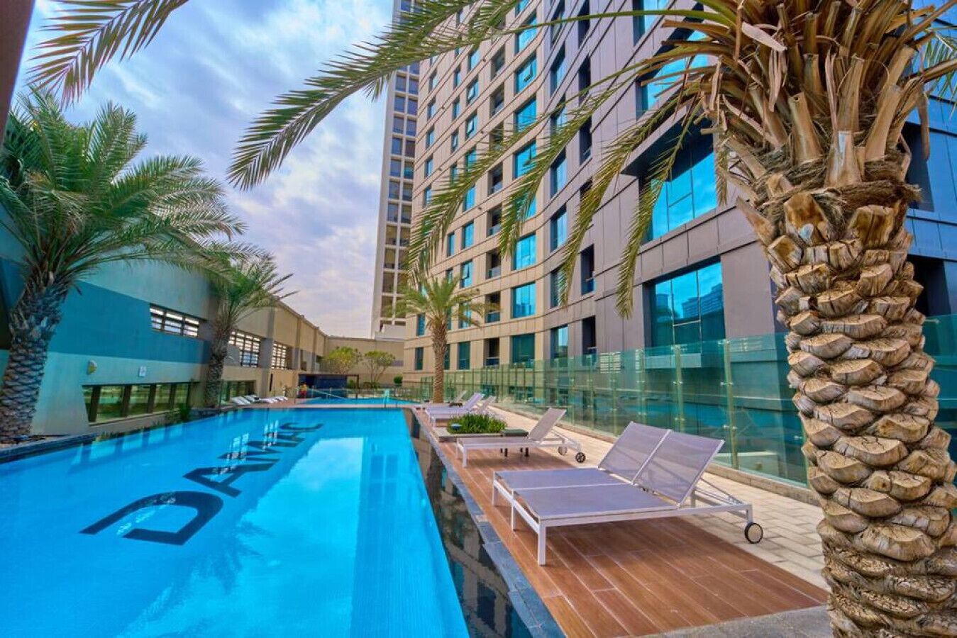 Un hotel moderno con piscina, palmeras y un rascacielos al fondo.