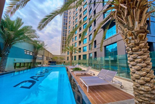 A modern hotel with a swimming pool, palm trees, and a high-rise building in the background.
