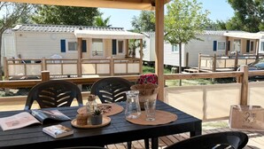 A wooden deck with a table set for dining, surrounded by chairs, and overlooking mobile homes. - ZEcamping 3* pataugeoire piscine locatif TV clim 24m2 4 pers (Saint-Mitre-les-Remparts)