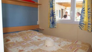A single bed with a floral bedspread and a straw hat on it. There is a window with curtains, and a bunk bed is visible in the background. - ZEcamping 3* pataugeoire piscine locatif TV clim 24m2 4 pers (Saint-Mitre-les-Remparts)