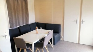 Ein kleiner Raum mit einem weißen Tisch, zwei Stühlen, einer Flasche und zwei Gläsern. Ein dunkel gefärbtes Sofa und eine Tür mit Griff sind ebenfalls vorhanden.