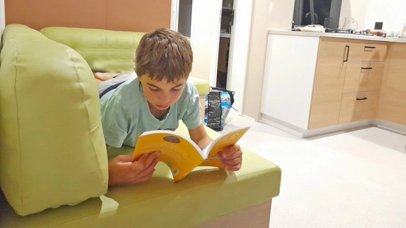 Una persona leyendo un libro en una habitación con un sofá verde, armarios de madera y una encimera blanca.