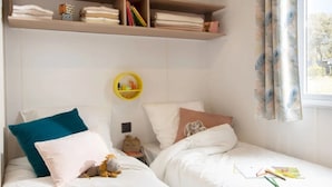 Une chambre avec deux lits, une étagère avec des serviettes pliées, une petite étagère jaune contenant des articles de toilette et une fenêtre avec des rideaux.