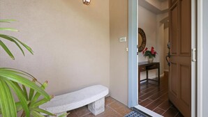 A hallway with a stone bench, a wall-mounted light fixture, a door with a number plate, and a potted plant. - Fairways at Mauna Lani Unit 1405 Garden View 3 Bed (Waimea)