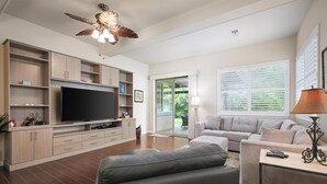 A modern living room with a flat-screen TV, wooden cabinets, a grey sofa, and a ceiling fan. - Fairways at Mauna Lani Unit 1405 Garden View 3 Bed (Waimea)