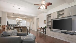 A modern living room with a sectional sofa, a flat-screen TV, and a dining area with a wooden table and chairs. - Fairways at Mauna Lani Unit 1405 Garden View 3 Bed (Waimea)