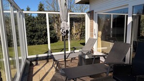 A covered patio with two lounge chairs, a table, and a glass door leading to a backyard.