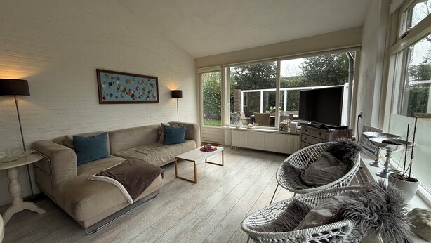 A spacious living room with a white brick wall, a beige sofa with blue cushions, a wicker chair, a white coffee table, and a television on a stand.