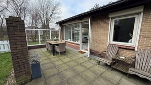 A patio with a wooden table and chairs, a brick wall, and a glass door leading to a garden.