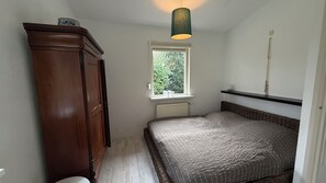 Ett litet, smalt rum med en trägarderob, en enkel säng med ett mönstrat överkast, ett fönster med utsikt över träd och en hängande lampa.
