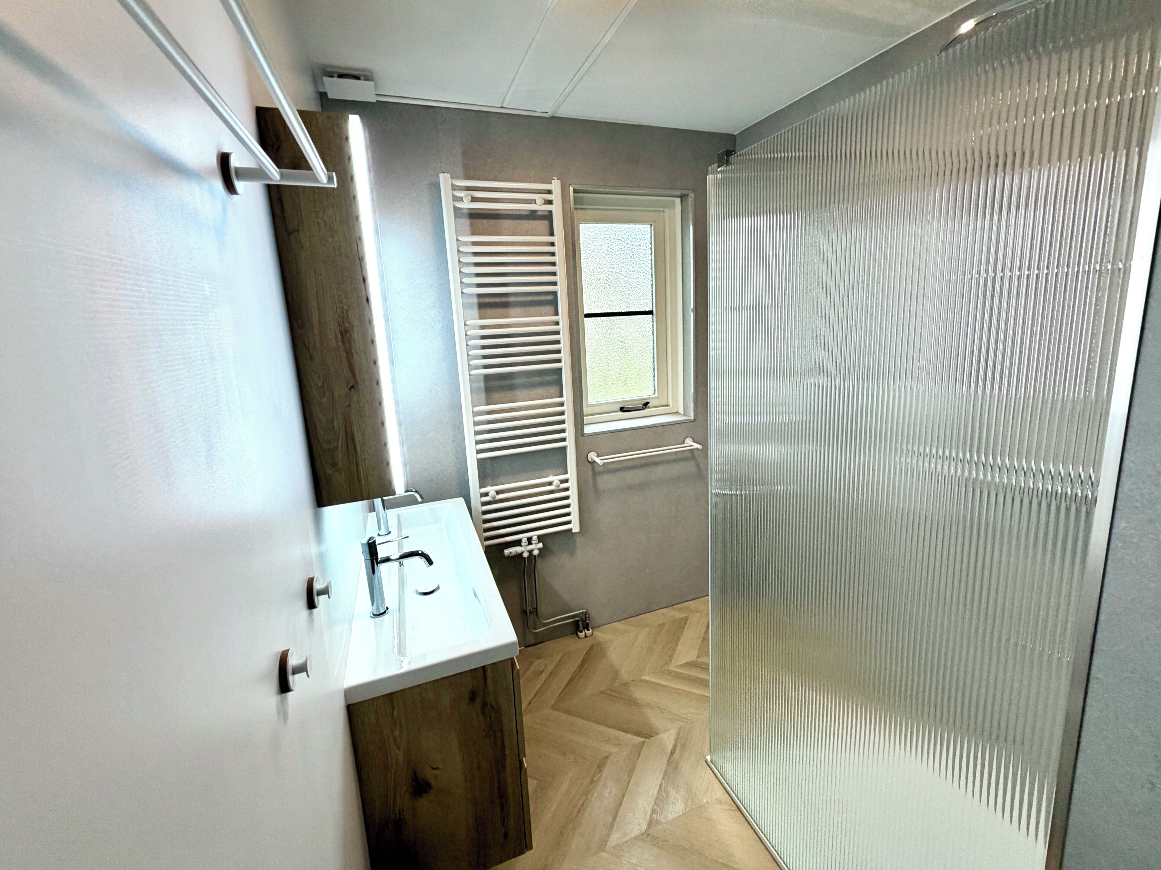 Een badkamer met een witte wastafel, een raam met handdoekhouder en een douche met een glazen deur.