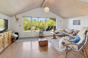 A cozy living room with a television, wooden cabinets, a wicker chair, and a large window offering a view of greenery. - Holiday home with peace, nature and great moments (Jægerspris)
