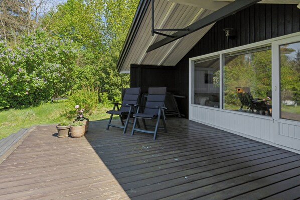 Eine Holzterrasse mit zwei Stühlen und Topfpflanzen, ein Gebäude mit großen Fenstern und Blick auf Bäume und Himmel.
