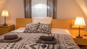 Un dormitorio con una cama, dos lámparas de noche, un cojín estampado y una toalla enrollada sobre la cama.